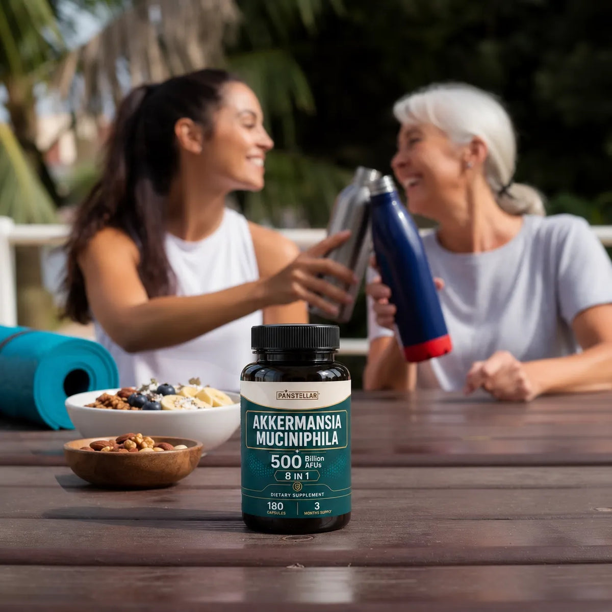 A bottle of Akkermansia Mucinipha 500 supplement is shown on a wooden table in a lifestyle setting, with two women smiling and laughing in the background while drinking from thermos bottles.