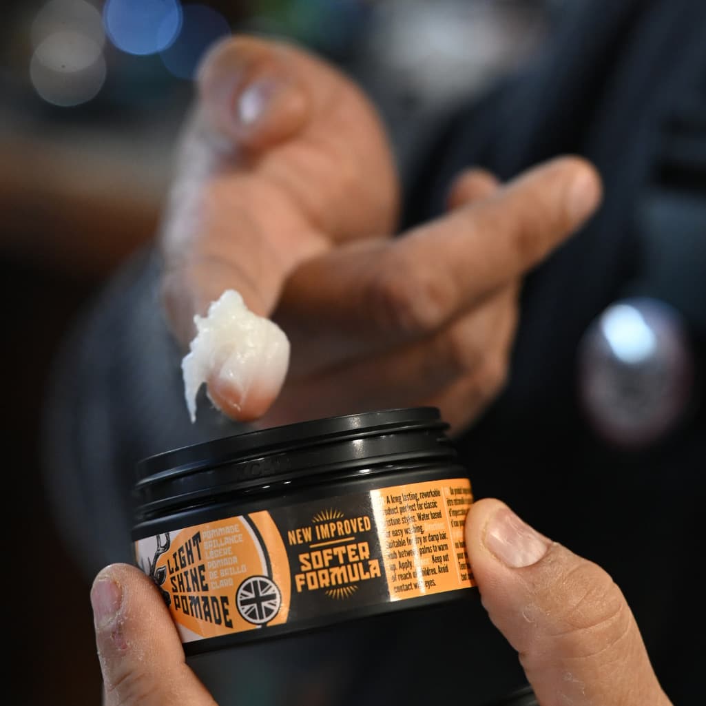 A close-up of a person holding a black jar of Softed Formula Bright Shine Pomade, with a dollop of the white pomade being spread on their finger against a dark, blurred background.