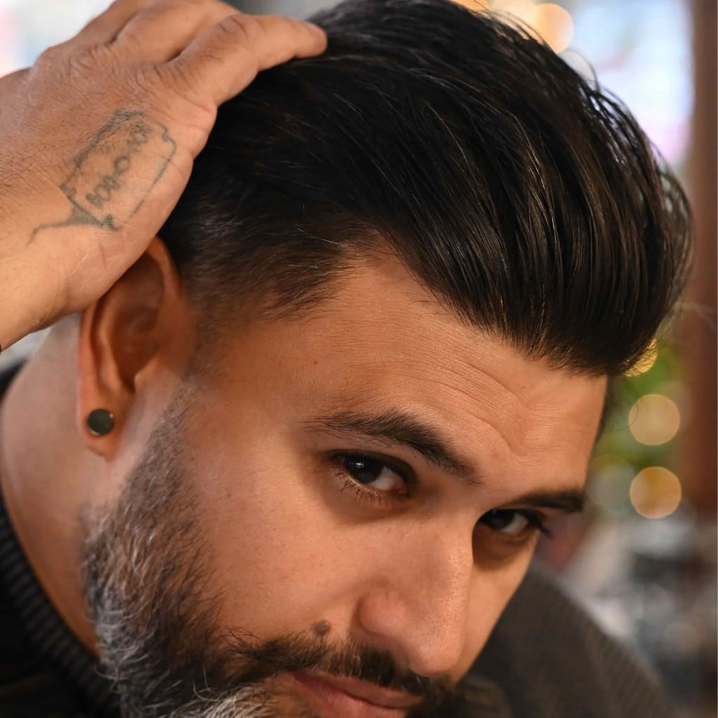 A close-up of a man with a styled dark brown pompadour hairstyle, showing the side and top of his hair. His hand rests on his head, and he has a beard and a small black ear cuff.