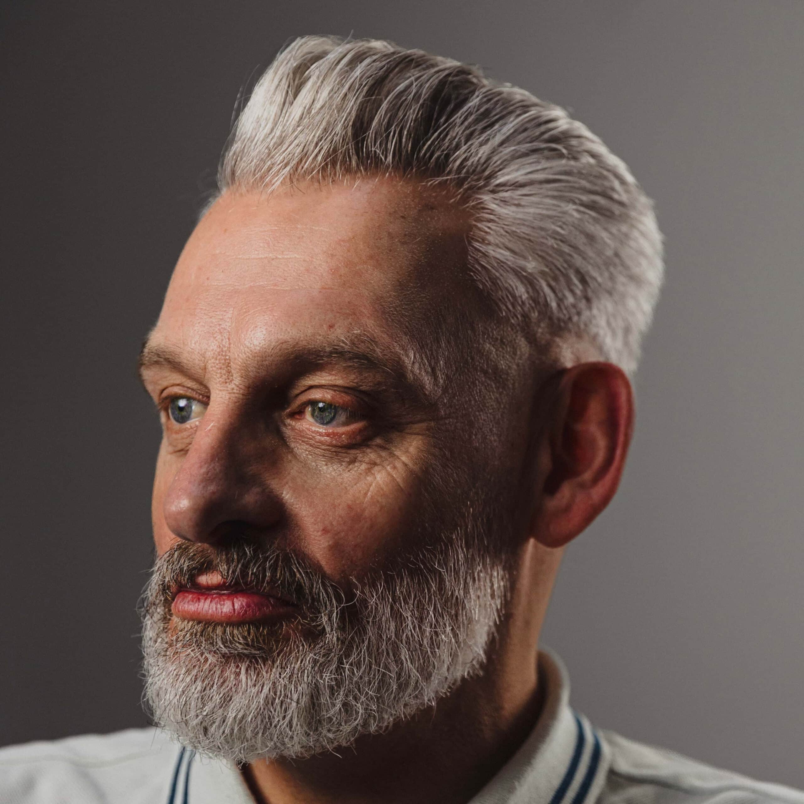A close-up portrait of a man with styled gray hair and a matching gray beard, wearing a light-colored collared shirt against a dark gray background.