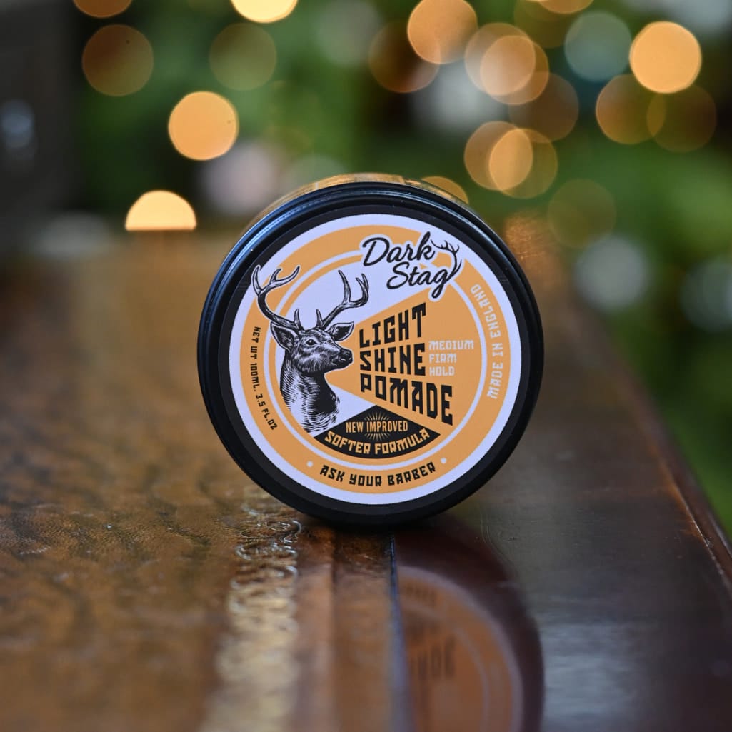 A round black container of Dark Stag Light Shine Pomade resting on a glossy wooden surface with a blurred background of warm bokeh lights.