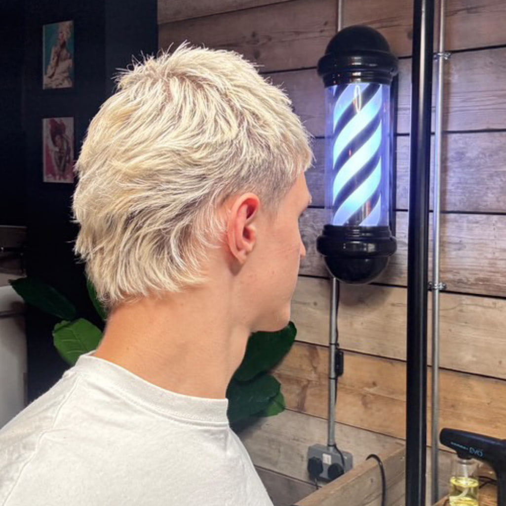 A man shown from the side profile in a barbershop setting, wearing a light gray t-shirt and displaying a short, textured blonde haircut. A classic black and blue barber pole is visible on the wooden-paneled wall behind him.