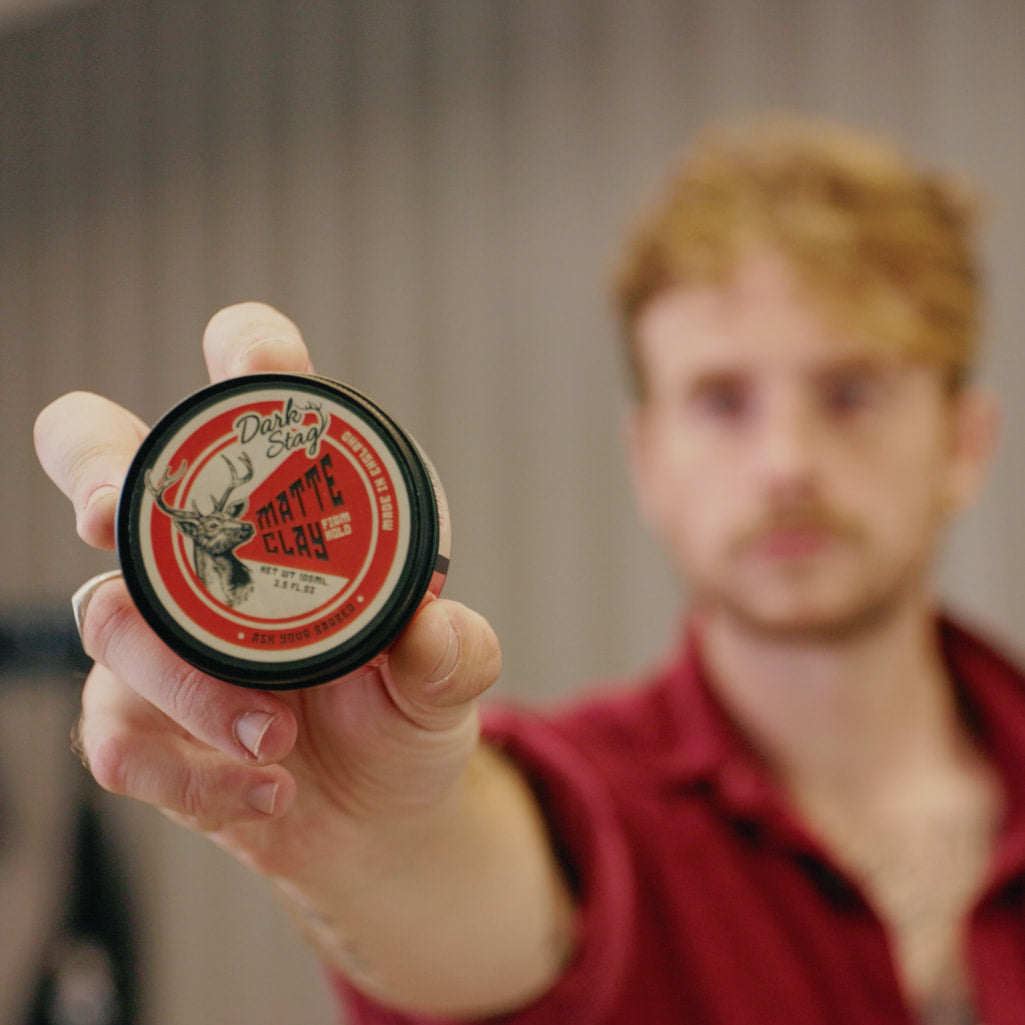 A man in a red shirt holds up a small round tin of Dark Star Matte Black hair wax toward the camera, with his face blurred in the background.