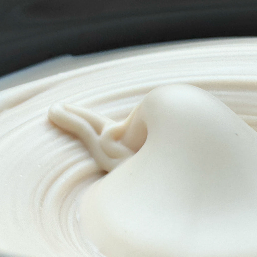 A close-up of a thick, creamy off-white cosmetic or food product, showing a smooth glossy texture with a swirled surface and a dark background.
