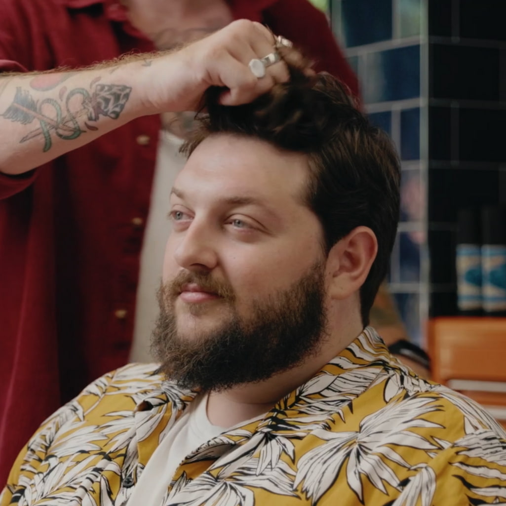 A man with a beard wearing a yellow and white tropical print shirt sits while another person in a red shirt styles his hair. The background shows a tiled wall and a wooden counter.