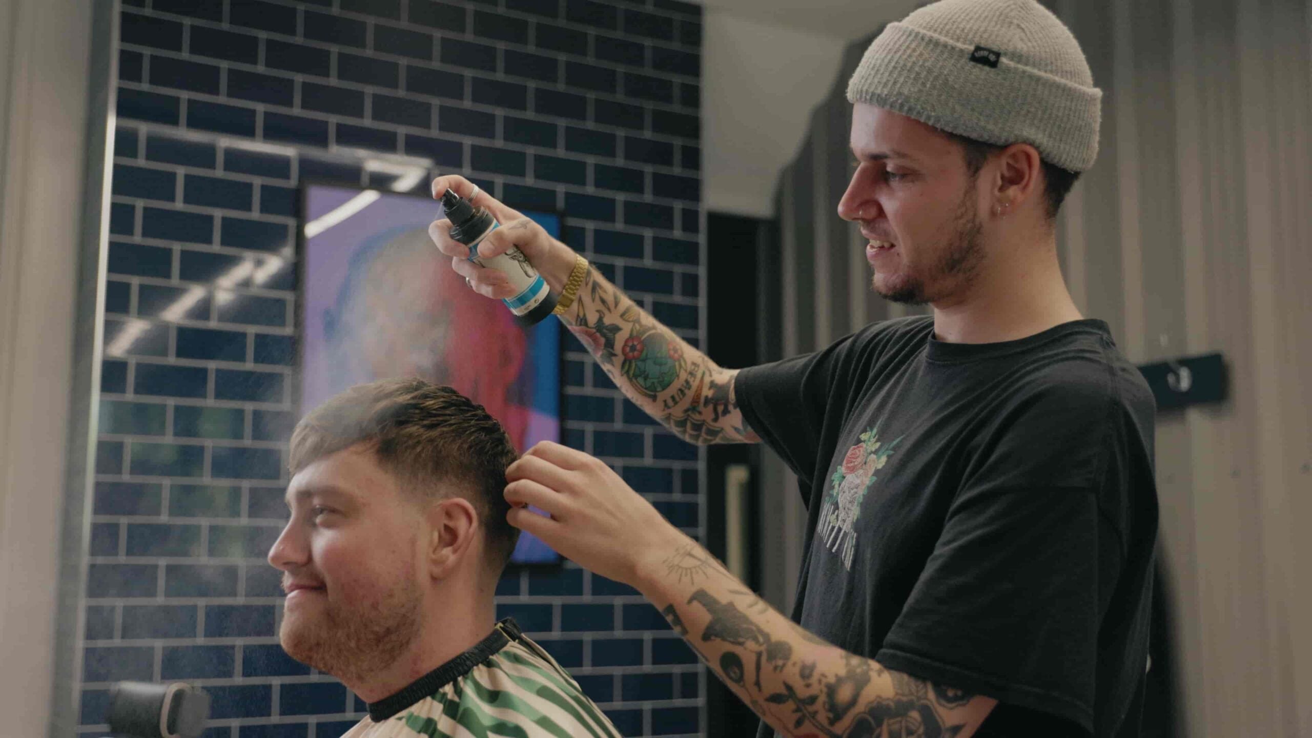 A man with tattoos wearing a gray beanie and black t-shirt sprays hair onto another man's head in a barbershop setting with a blue brick wall background.