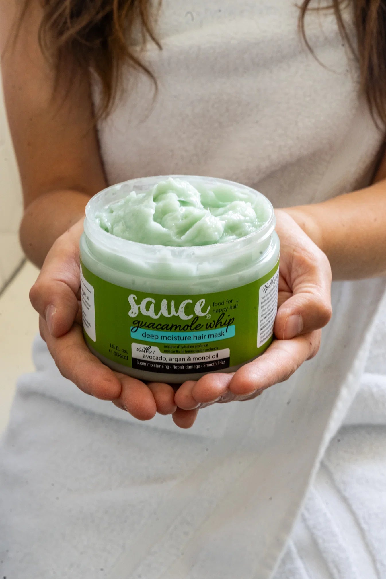 A person holding a jar of Sauce Guacamole Deep Moisture Hair Mask, showing the light green product inside a clear jar with a green label.