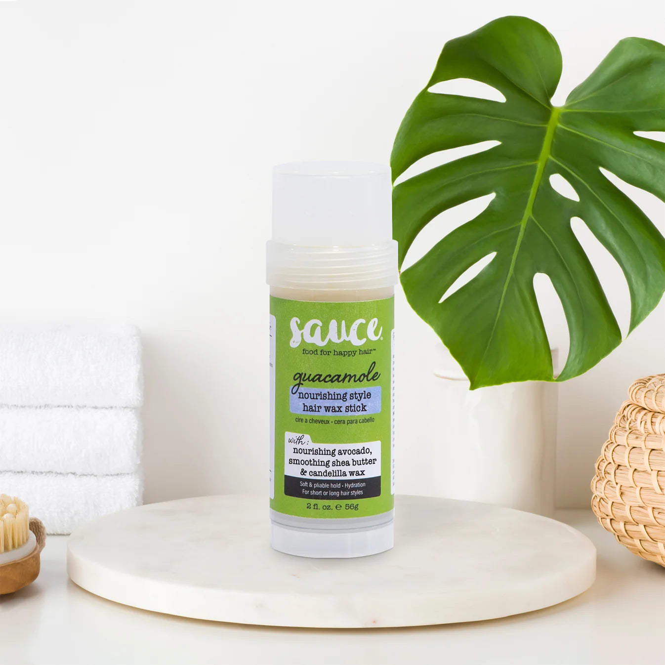 A bottle of Sauce Beauty Guacamole nourishing style hair wax stick stands on a round white marble tray. The product is styled in a bright, clean setting with a large green monstera leaf, white towels, and a woven basket in the background.