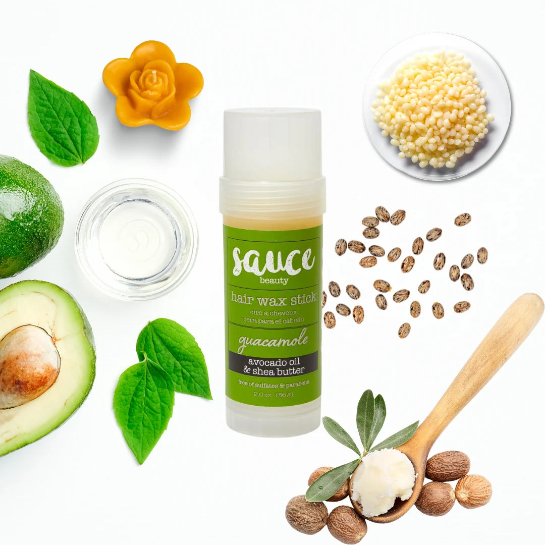 A bottle of Sauce Beauty Hair Wax Stick in guacamole, surrounded by its natural ingredients including avocado, green leaves, a yellow flower, chia seeds, and a wooden spoon with nut butter, all on a clean white background.