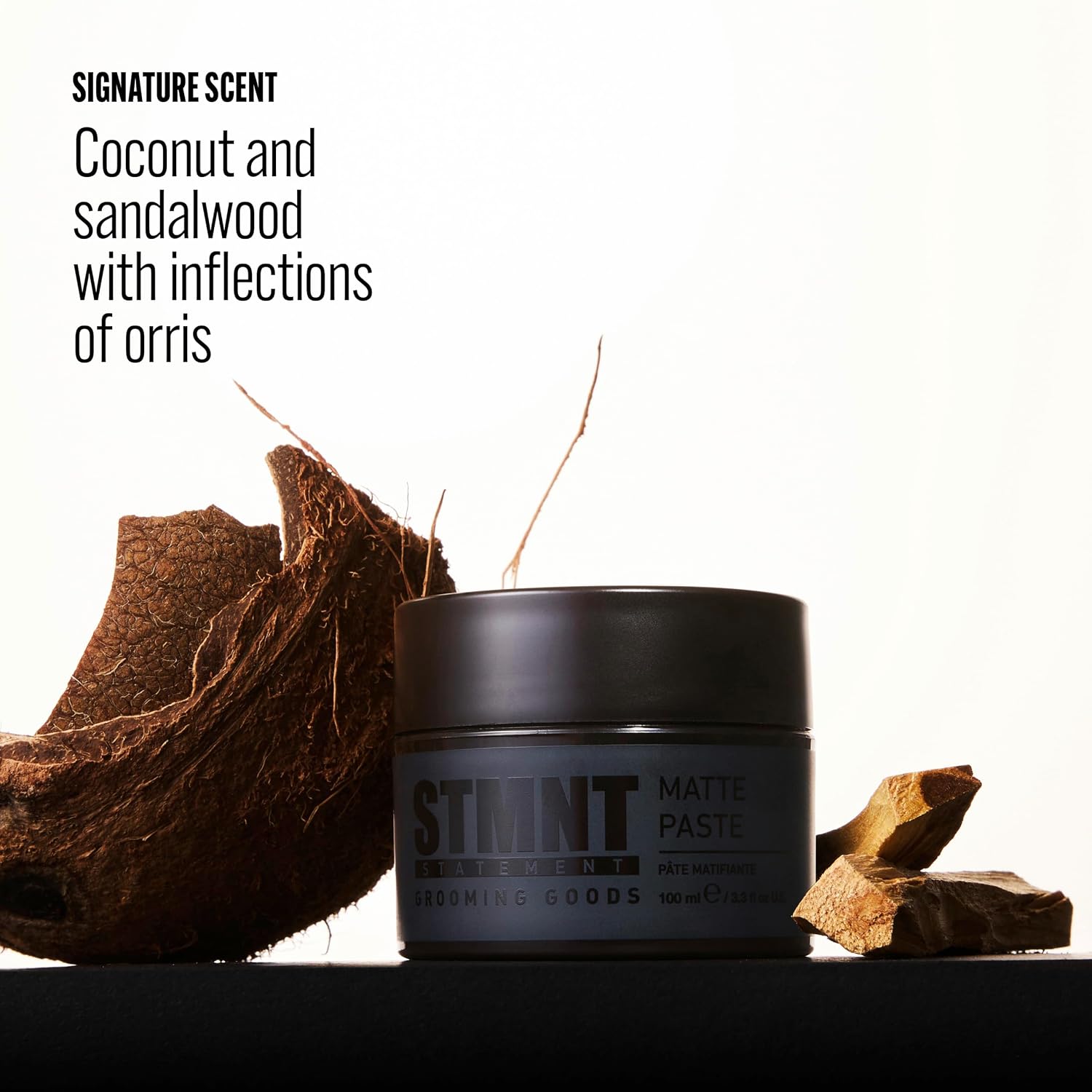 A dark gray jar of Stimnt Cosmetics Matte Paste hair product sits on a black surface with a coconut and wood pieces beside it, against a white background with text describing the scent.