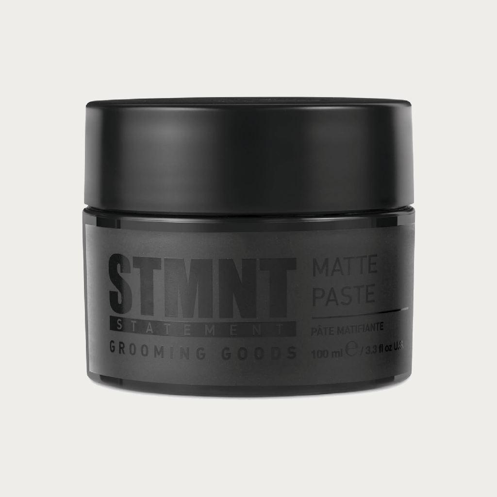 A black cylindrical jar of STMNT Statement Grooming Goods Matte Paste, shown against a light off-white background.